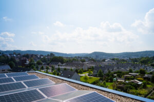 Eine Aussicht auf Wuppertal. In der Ferne sind Häuser, Berge und die Sonne scheint. Im unteren Bildbereich sind Photovoltaik.
